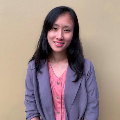 Mindy is facing the camera smiling, wearing a lavender dress coat over a pink blouse. Behind her is a cream-colored, blank wall.