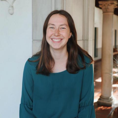 Catherine is standing in the middle frame, facing the camer and smiling. She is wearing a green blouse. Behind her is a white doorframe, and in the back you can see an ionic column.