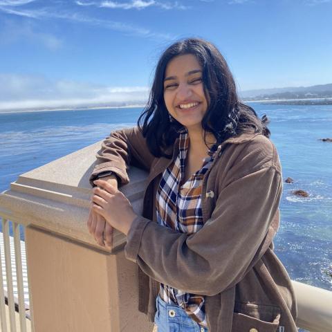 Saadhana is seen facing the camera, smiling. They are wearing a beige coat over a plaid-patterned buttoned shirt loosely tucked into light-wash jeans. They are leaning on a post. Behind them is the ocean with some light clouds in the sky.