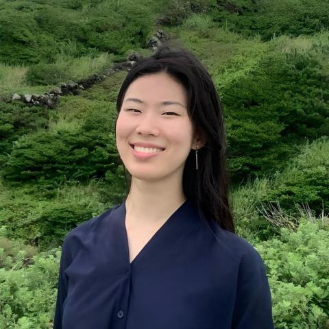 Hannah is standing in the middle of the frame, facing the camera and smiling. Behind her is green shrubbery.