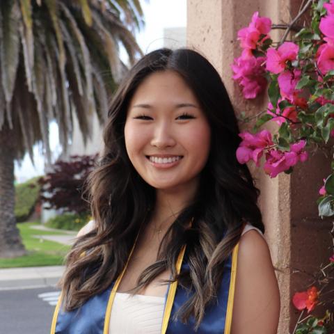 Megan Li is facing the camera and smiling. She is wearing a white dress. She is wearing a blue and gold graduation stole over her shouldes. Flowers and a palm tree frame the background. 