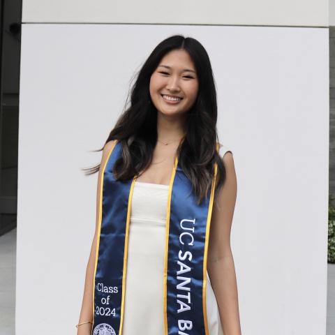 Megan Li is facing the camera and smiling. She is wearing a white dress. She is wearing a blue and gold graduation stole over her shouldes. 