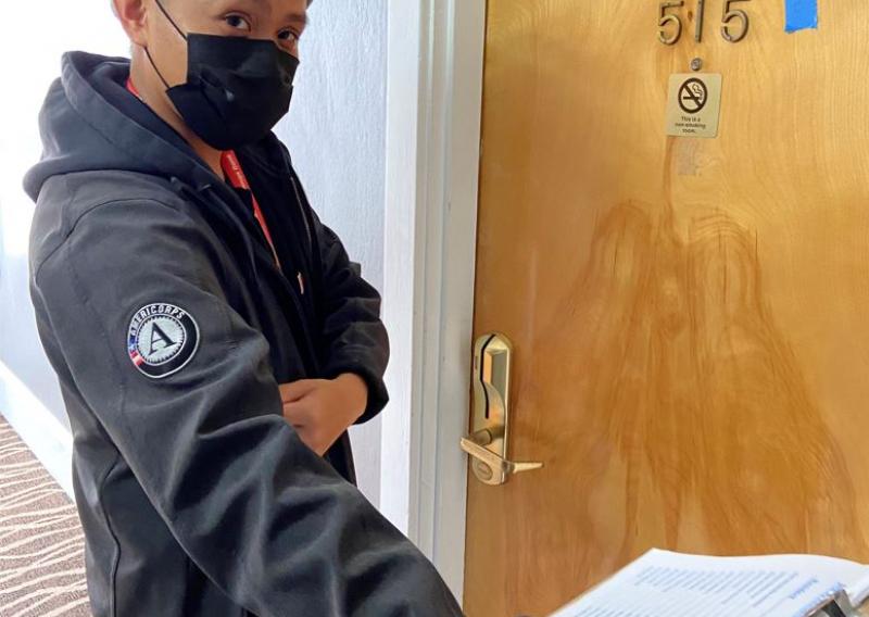 Jeffrey Levita facing the camera with a facemask on. He is extending his right arm out to the lower right corner of the frame while holding a clipboard. On his right shoulder is the AmeriCorps logo.