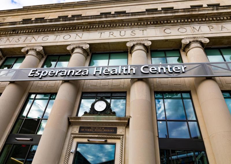 Looking up at a building with tan columns. There is an ornate clock over the front door and above that a sign that says "Esperanza Health Center"