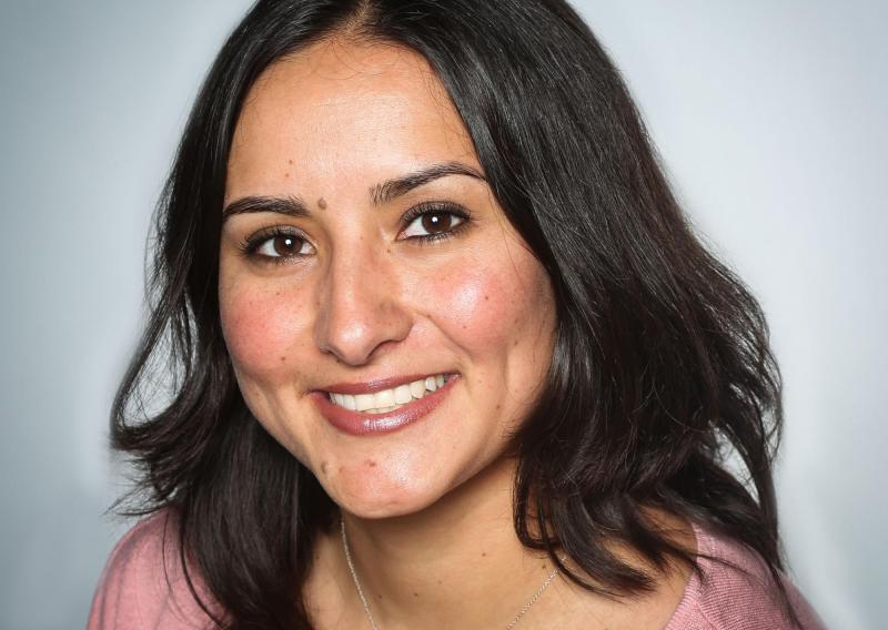 Lily Morales is seen here facing the camera and smiling. Her photo was taken against a grey white background. She is wearing a wide neck pink blouse.