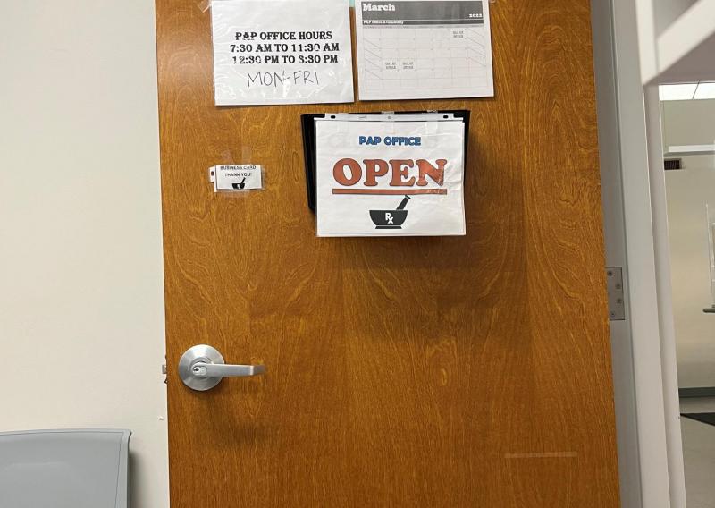 An open door with four signs hanging. At the top a sign reads "PAP" under that are two, on the left lists the office hours and on the right is a calendar showing March. The last sign is below all of them and it reads "PAP Office Open" 