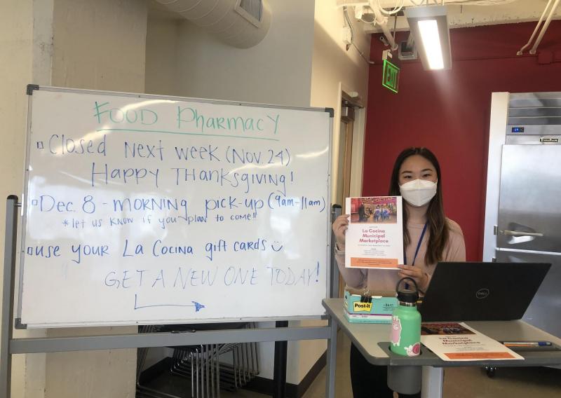 Joelle is holding a flyer for La Cocina's Municipal Marketplace. Food Pharmacy's whiteboard indicating information regarding Thanksgiving closure, a December food pharmacy pickup, and La Cocina giftcards is to her left.