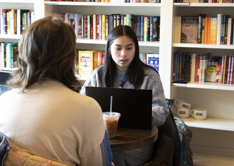 Patricia sitting in front of a bookshelf with her laptop. There is another person sitting across from her whose face we cannot see.