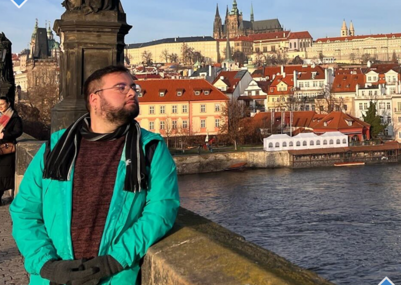 Alan is standing to the left of the camera frame, looking to their left. They are standing on a bridge, and behind them is the Prague skyline. 