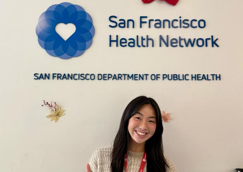 Sam stands in the center of the frame, facing the camera and smiling. Behind her is the logo for the San Francisco Health Network.