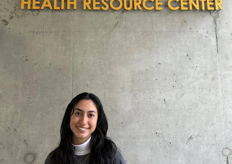 Laura is standing off center frame, facing the camera and smiling. Above her is the entrance sign for the Maria X Martinez Health Resource Center.