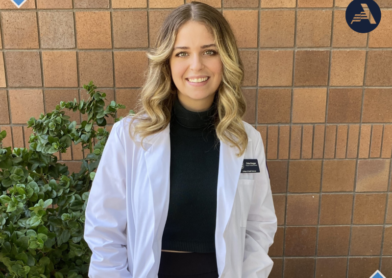 Chloe Reagan stands in the middle of the frame, facing the camera and smiling. Chloe is wearing a white coat over a black turtleneck. 