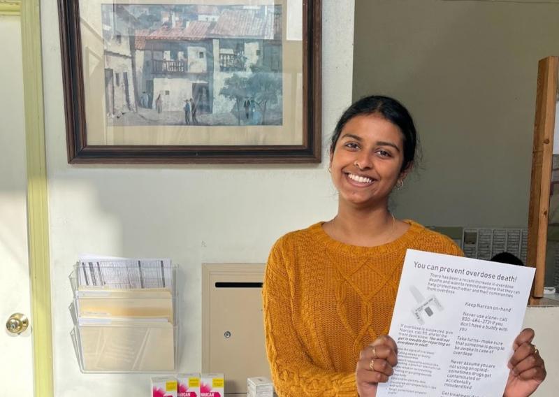 Nivedha stands in the off-right of the frame, facing the camera and smiling while holding a health resoruces flyer.