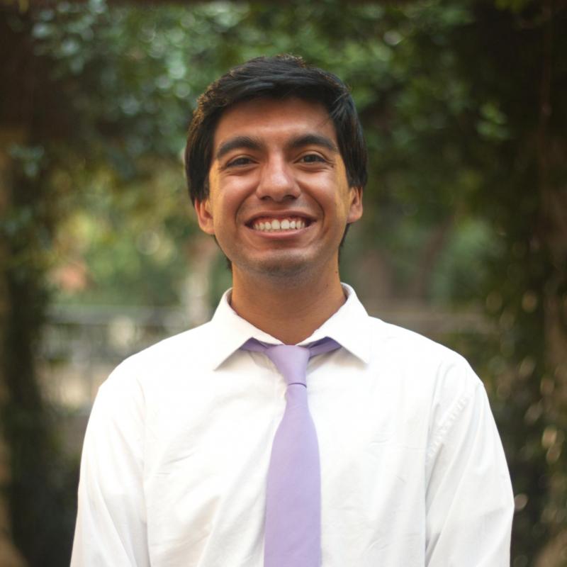 Jonathan Reyes is facing the camera, smiling, wearing a white button up shirt and a lavender tie. The background is composed of two trees on both sides of Jonathan.