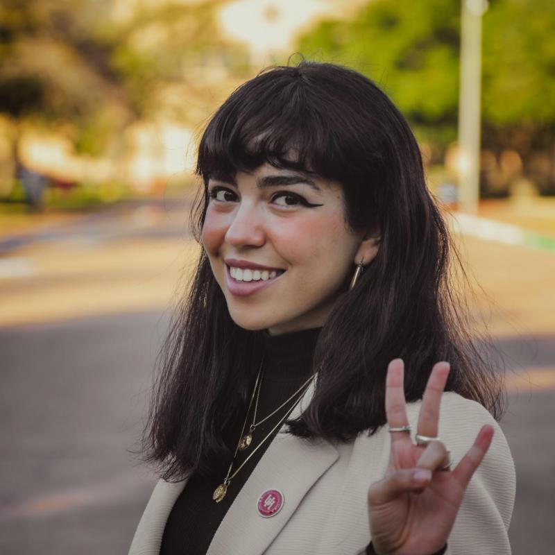 Jasmine is in the center of the frame, is facing slightly to her right. She is making a peace sign in the photo. She is wearing a tan blazer and a black turtle neck. 