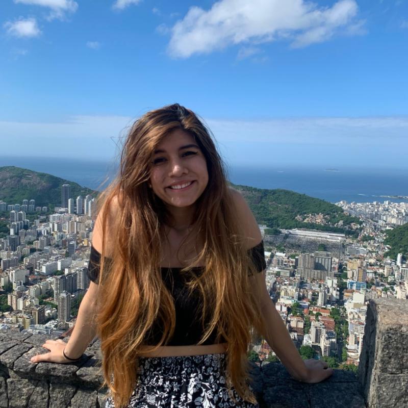 Paola is standing in the middle of the frame, leaned against a low stone railing and smiling at the camera. Behind her is a view of a city, rolling hills, and the ocean in the distance. 