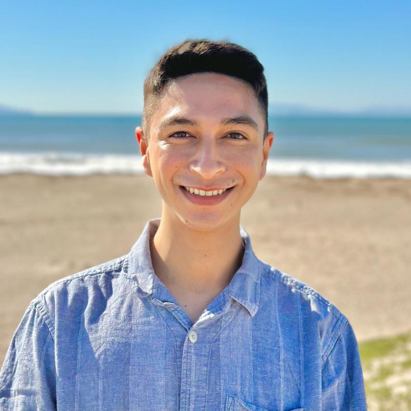 Cameron is standing in the middle of the frame, smiling at the camera. Behind him is a beach, with the sand followed by the sea in the distance. 