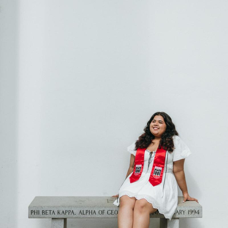 NHC member Ann Sunny wearing a white dress and graduation stole, sitting on a bench.