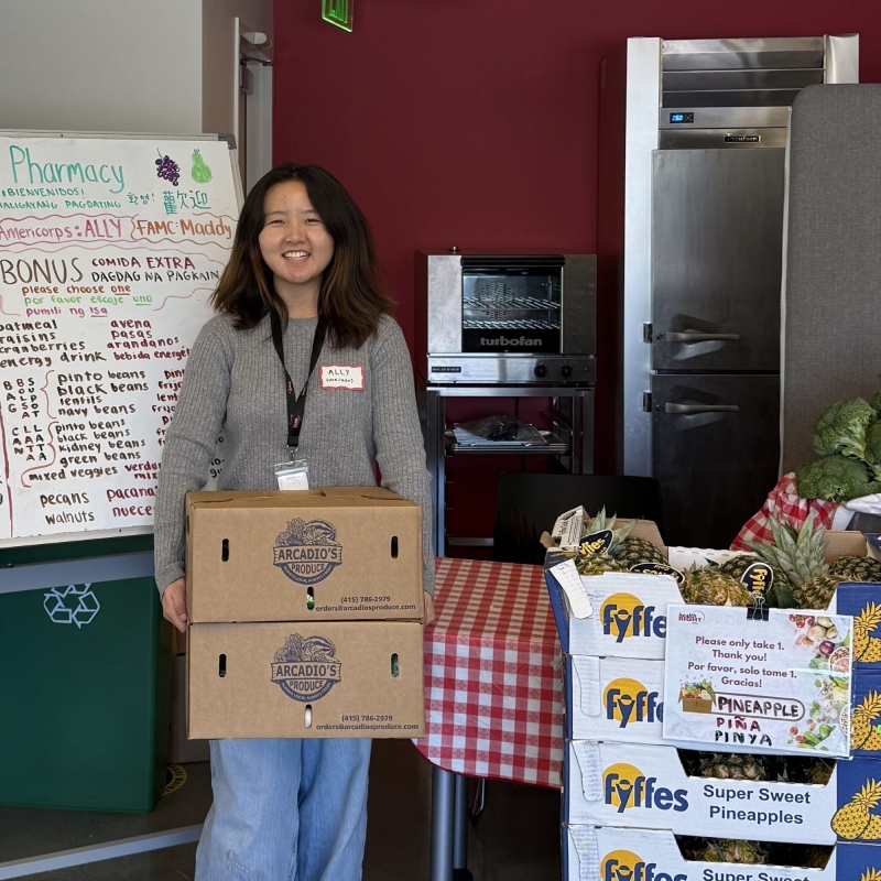 Ally is standing to the left of the frame holding two boxes of produce. Behind her is a white board explaining what is in each box, and to the right of her are stacked boxes filled with a variety of vegetables.