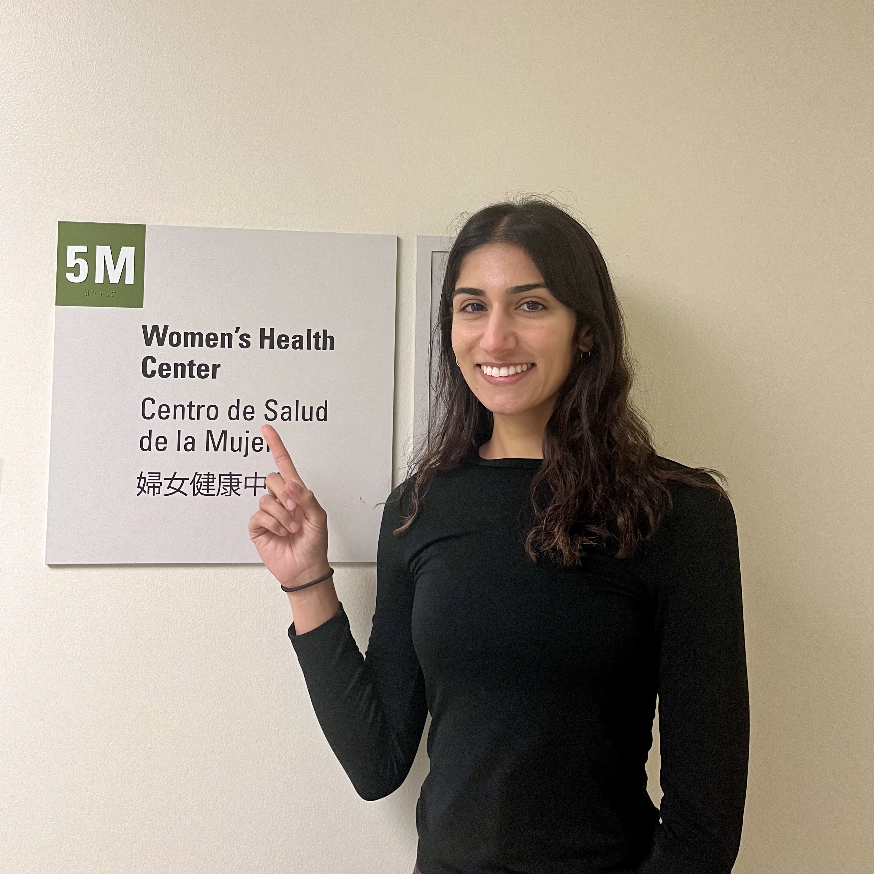 Anisha is seen in the center of the frame, facing the camera and smiling. She is pointing at a sign that reads "5M: Women's Health Center." The sign is also translated into Spanish and Chinese.