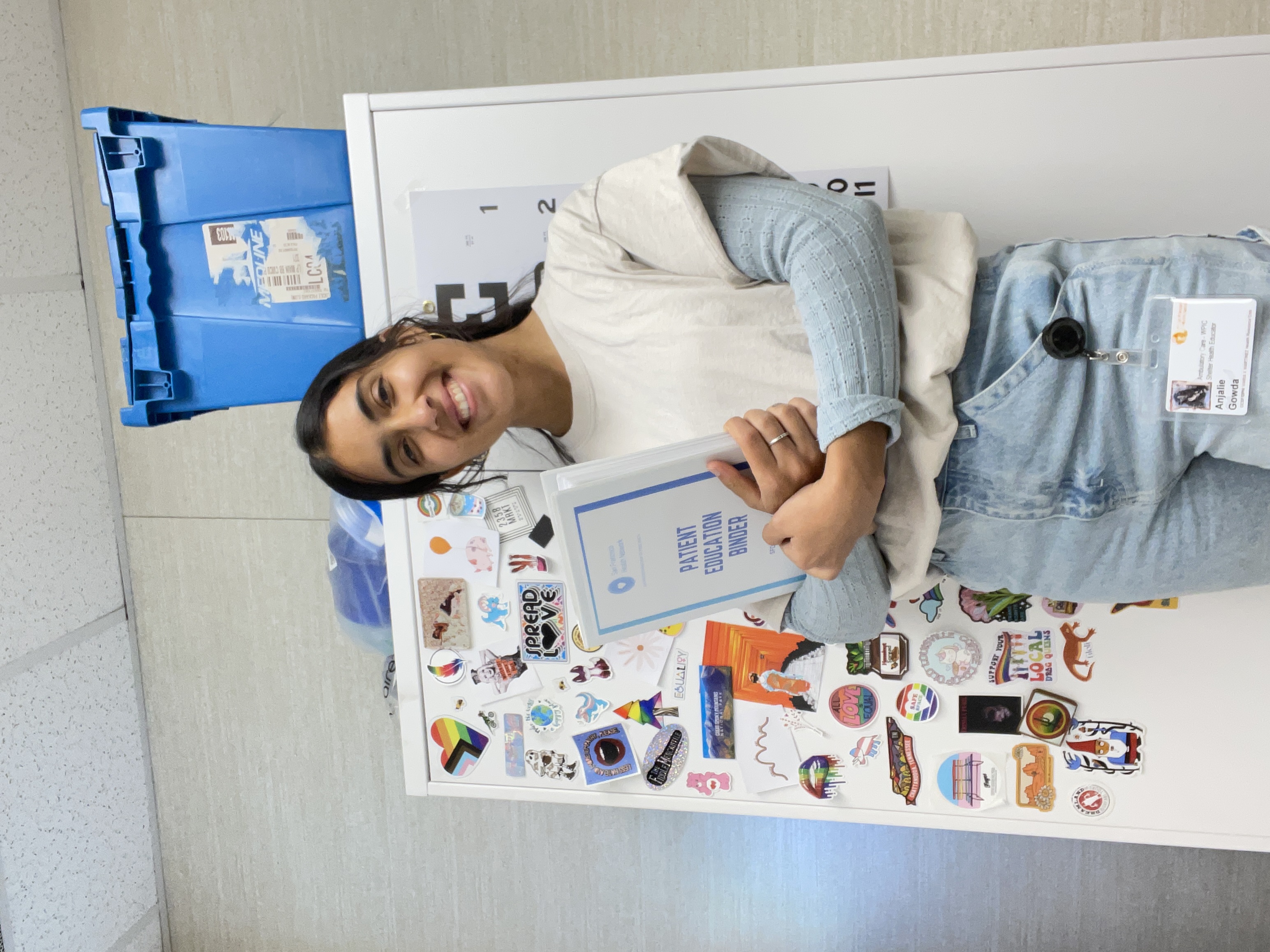 Anjalie is standing in the center of the frame, holding a binder. The cover of the binder reads "Patient Education Binder."