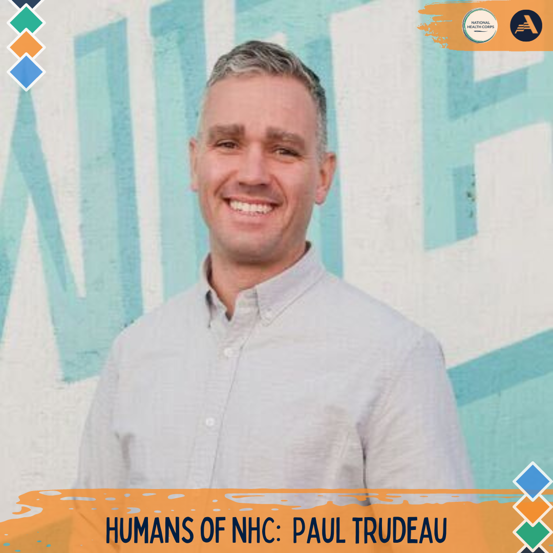 Paul Trudeau stands in the middle of the frame, facing the camera and smiling. He is wearing a white, collared button up shirt. 