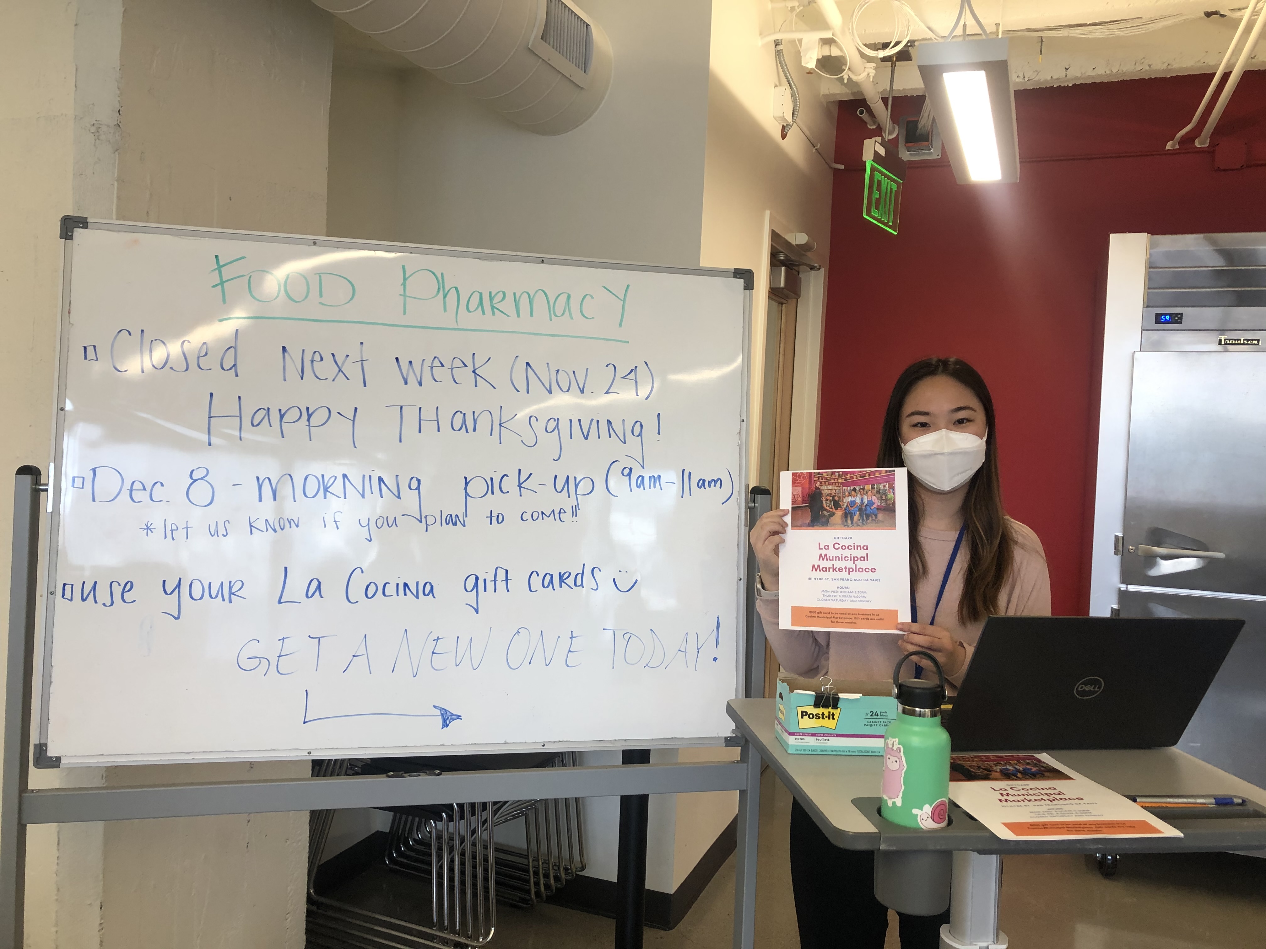 Joelle is holding a flyer for La Cocina's Municipal Marketplace. Food Pharmacy's whiteboard indicating information regarding Thanksgiving closure, a December food pharmacy pickup, and La Cocina giftcards is to her left.