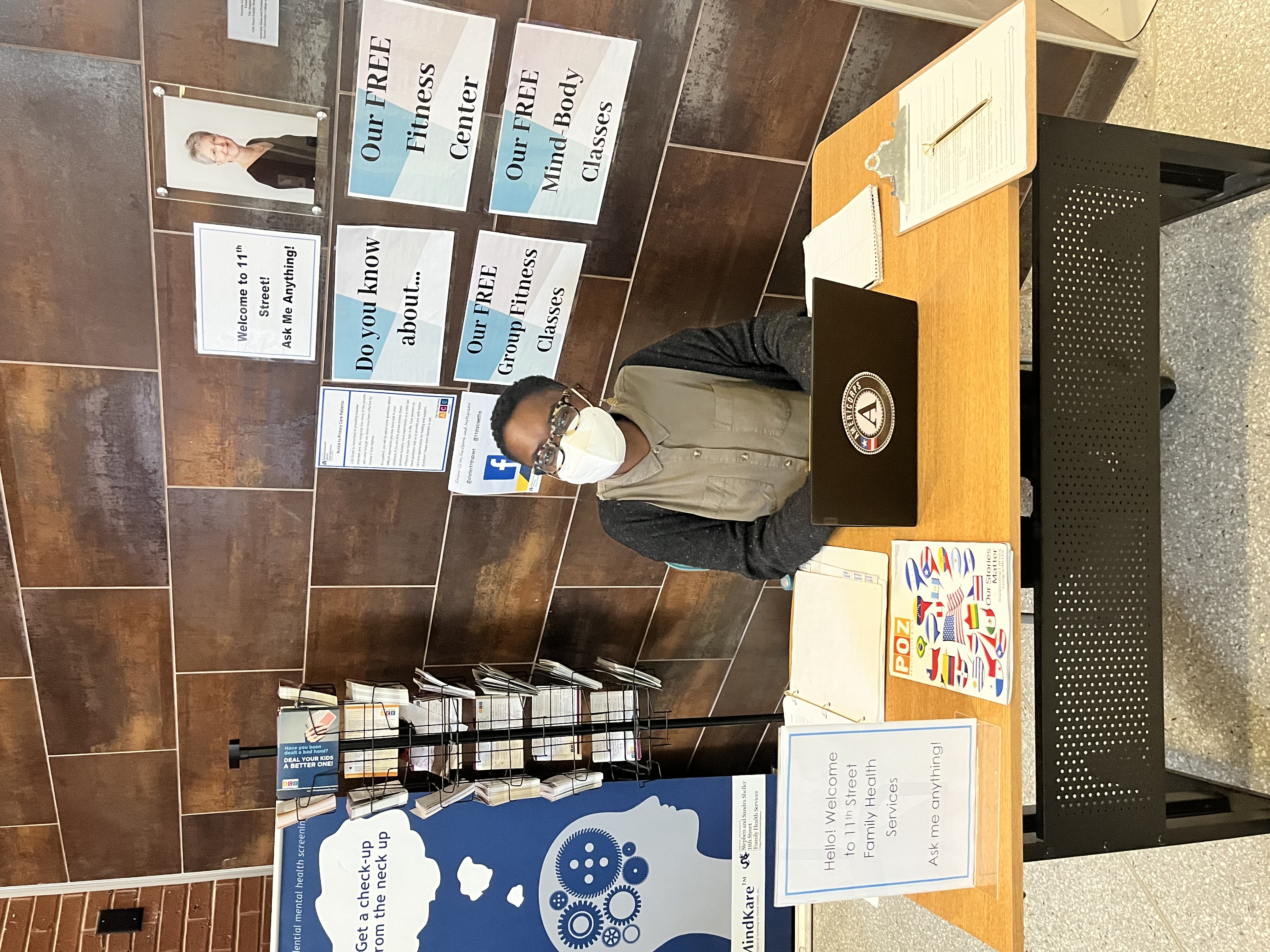 Jessica sitting at a table with a laptop and a sign that says "Hello welcome to 11th Street Family Health Services, Ask me anything" behind her are signs advertising group fitness classes and mind body classes