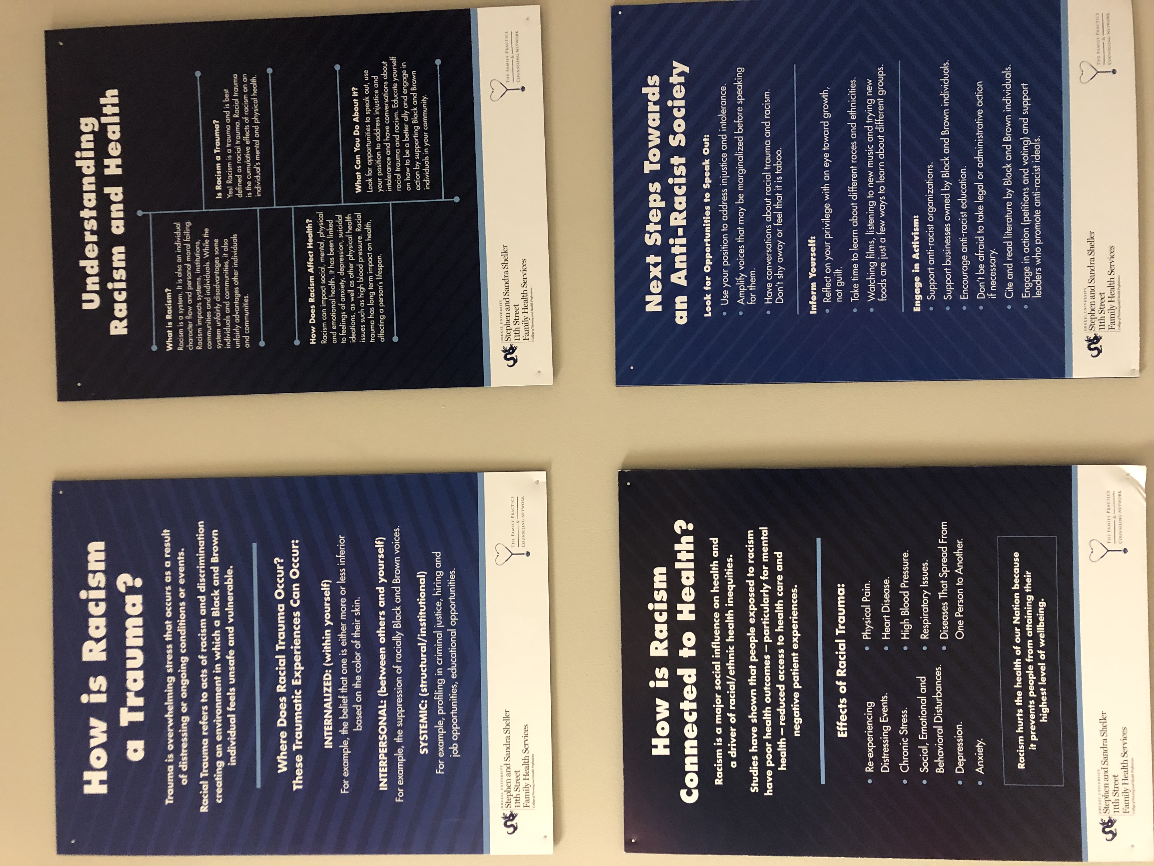 Four blue signs hung on a wall. Clockwise from top left hey are titled "How is Racism a Trauma" "Understanding Racism and health" "How is Racism connected to health" and "Next Steps Toward an Anti Racist Society"