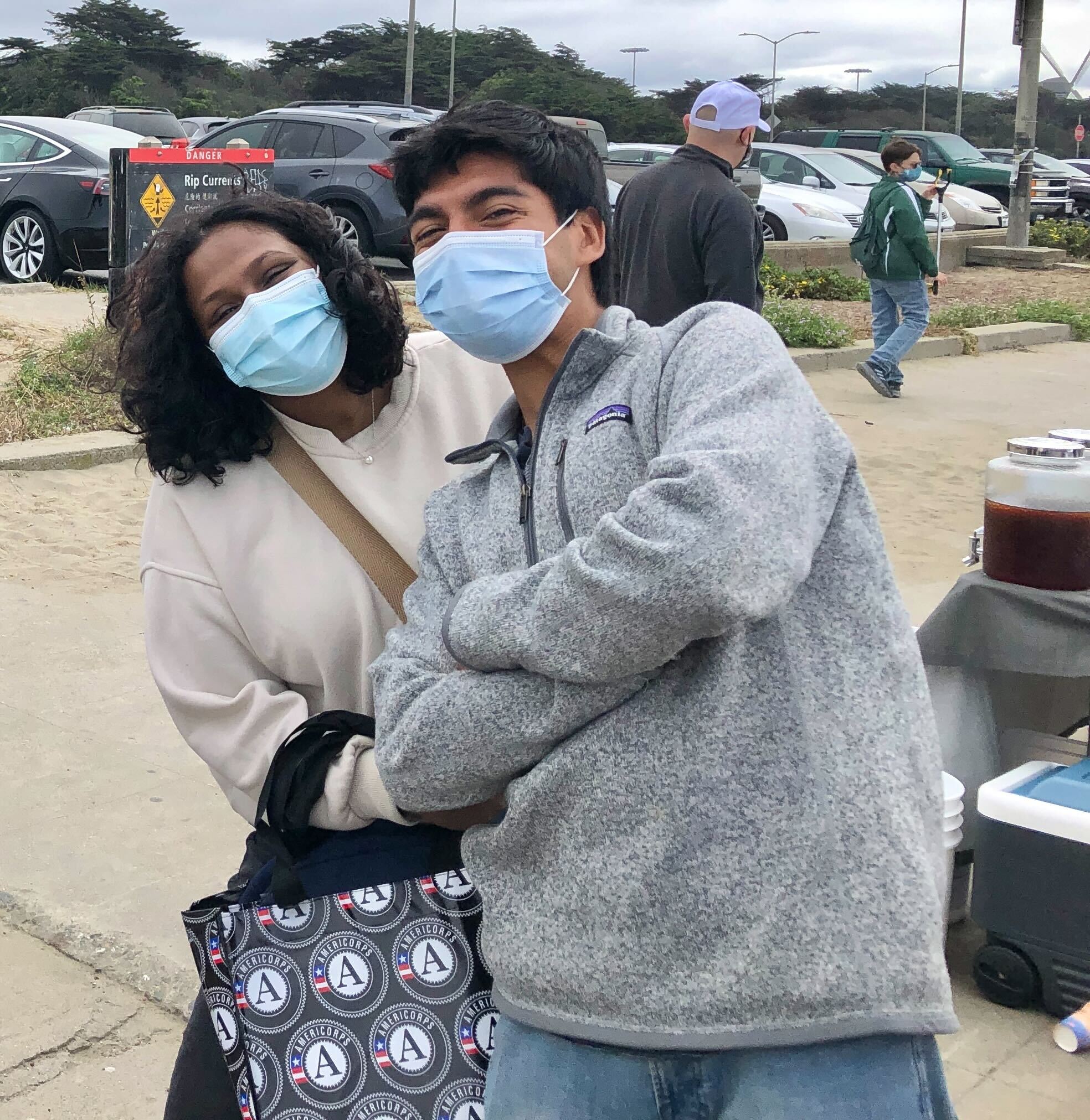 Two people are in the center of the photo. Johnny Reyes (right) and Sneha Suresh (left) are leaning into each other, arms crossed in front of their bodies. They both have a facemask on.