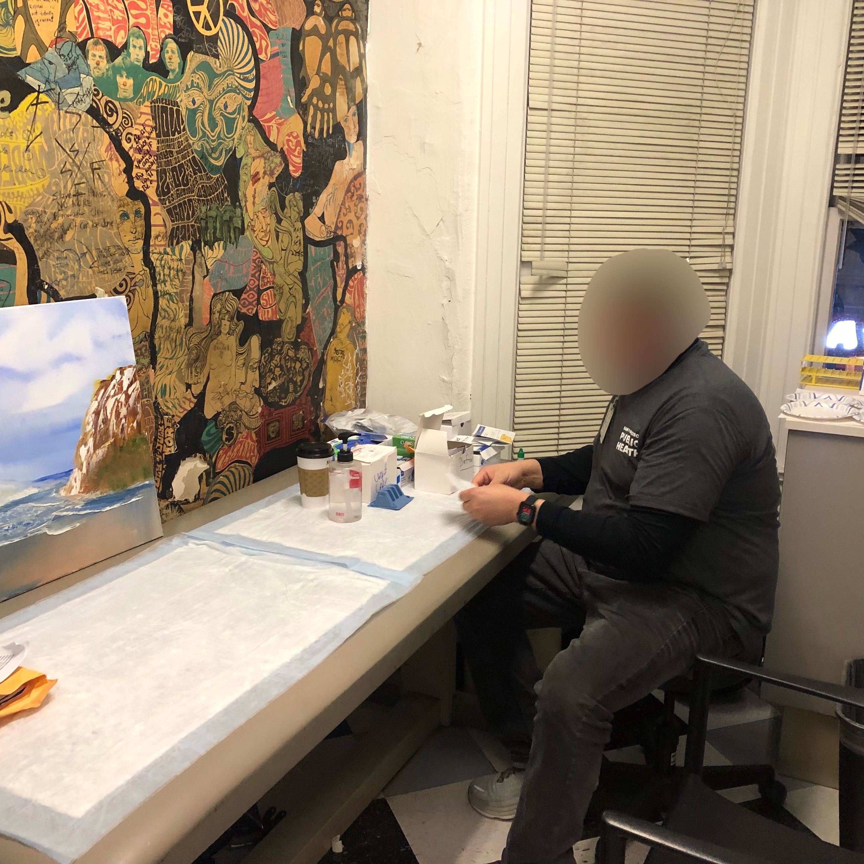 A health care worker (face blurred) is sitting at a testing table preparing the table to conduct multiple health exams.