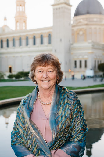 A person is standing at the center of the photo and is seen from the waist up. They have their arms crossed in front of them and are smiling at the camera. In the background there is a tall building.