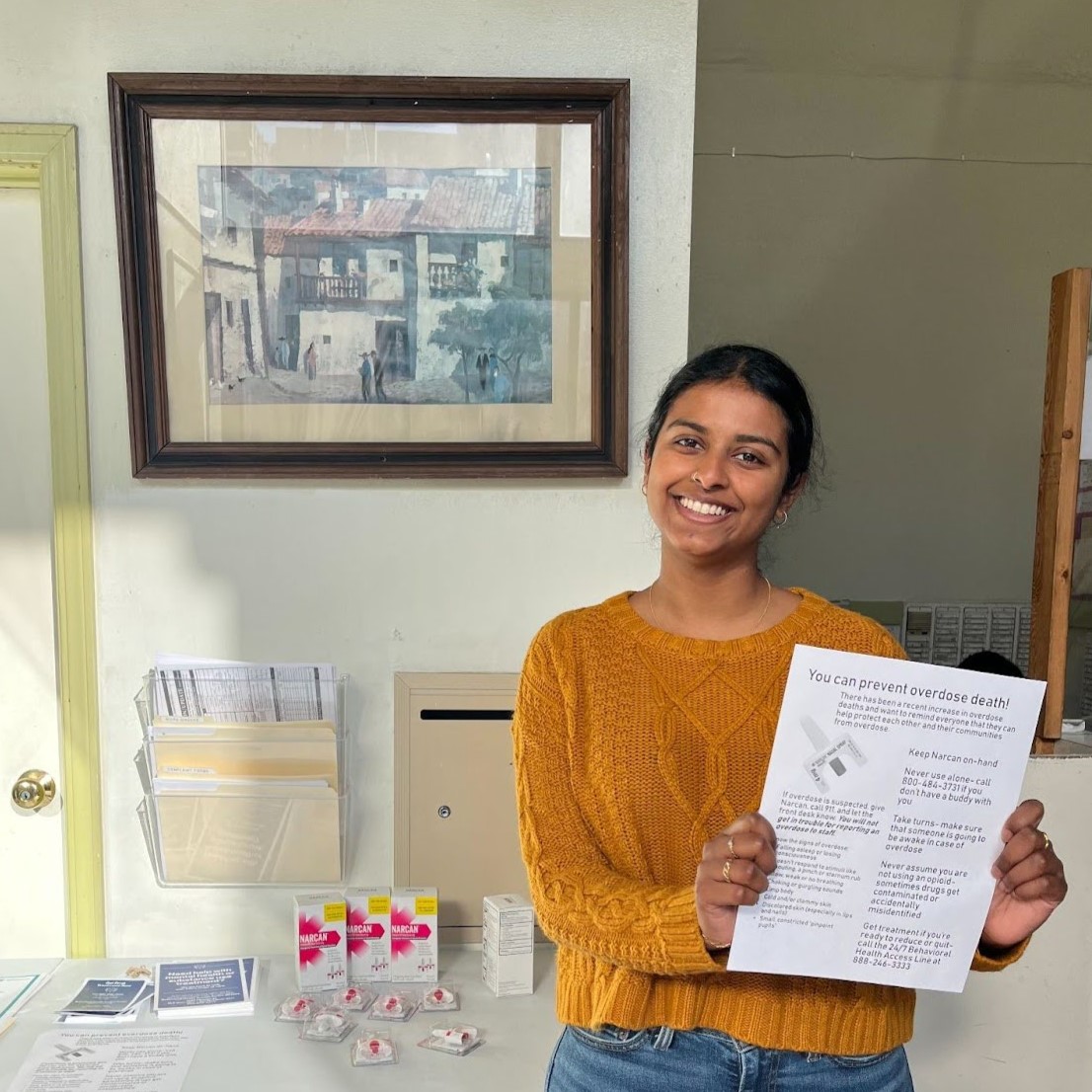 Nivedha stands in the off-right of the frame, facing the camera and smiling while holding a health resoruces flyer.