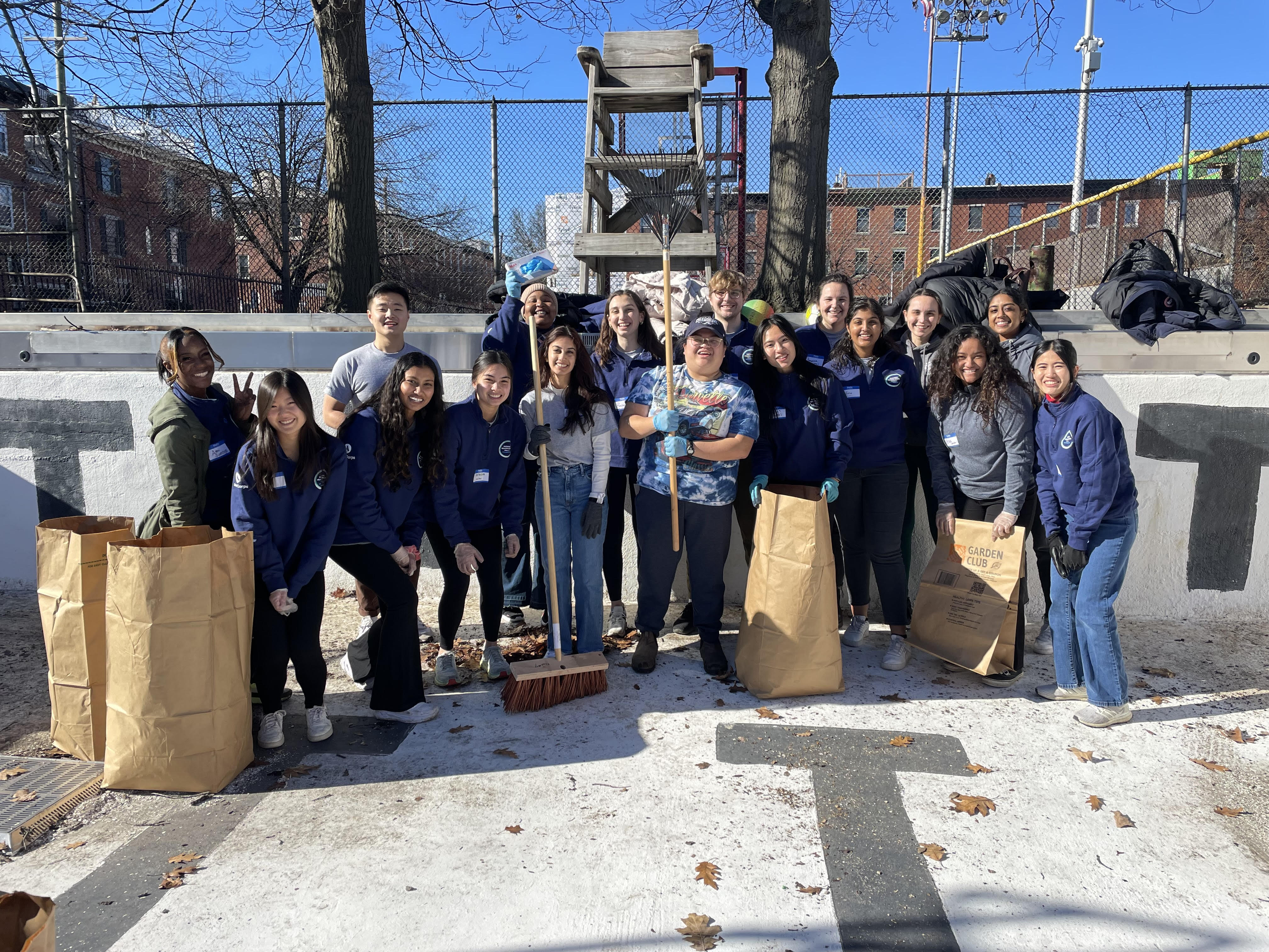 A group photo of NHC members holding brown paper yard waste bags, brooms, and rakes while standing in an empty pool clearing out leaves